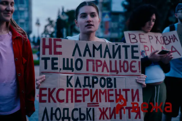 Two young protesters hold cardboard signs with red and black Cyrillic text at a demonstration, the central woman speaking toward the camera as dusk settles behind them.