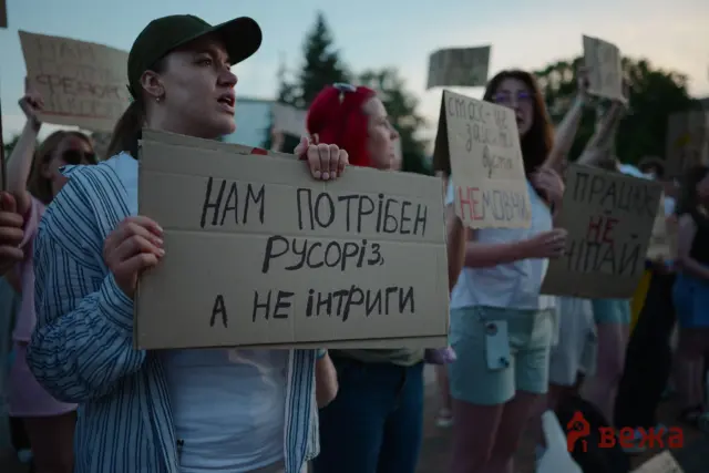 Protesters hold cardboard signs at a rally; woman in a striped shirt and green cap foreground, others behind with handwritten signs in Cyrillic.
