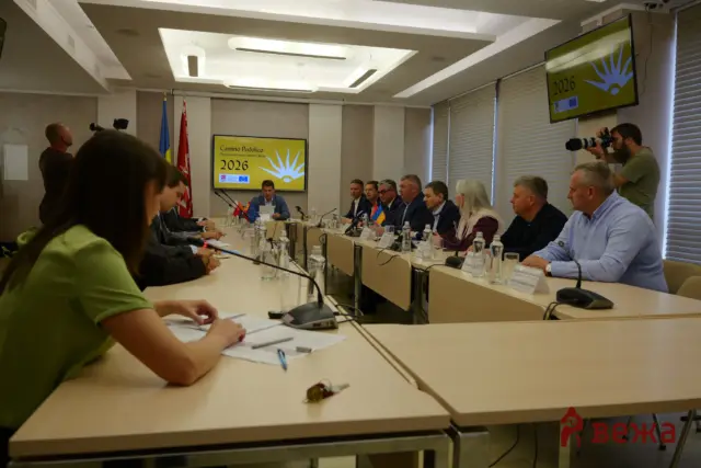 Officials sit around a long conference table in a meeting room; a photographer with a camera stands at right, flags and screens display '2026' and event branding.