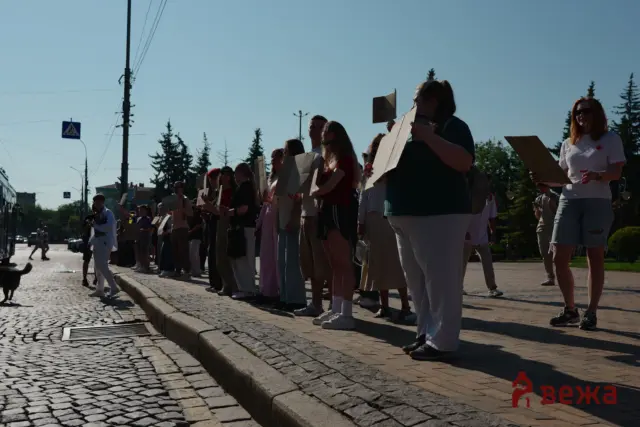 A line of protesters on a sunny street holding cardboard signs along a cobblestone sidewalk.