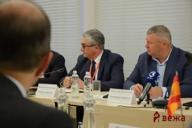 Panel of three suited men in a conference room, seated at a table with nameplates, microphones, and water bottles; foreground person partially visible from the back.