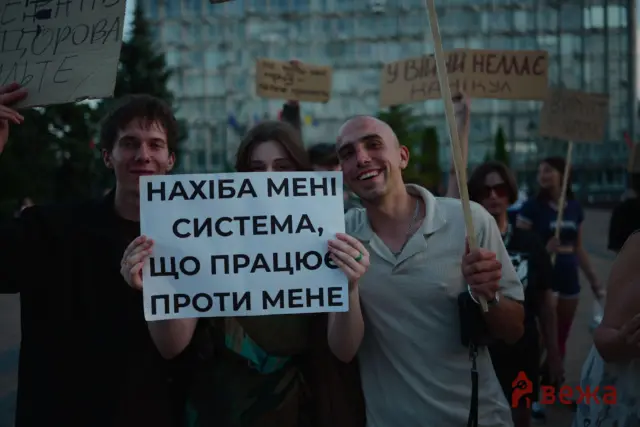 Protesters at a rally holding signs, with a large sign in Ukrainian reading 'НАХІБА МЕНІ СИСТЕМА', smiling faces in the foreground and an urban background.