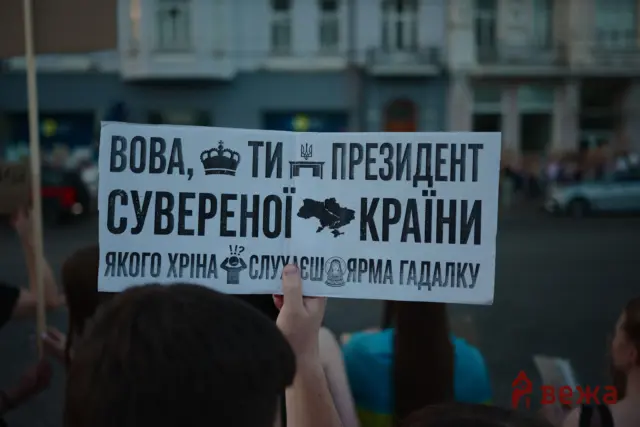 Protest sign with large Cyrillic text and a small map of Ukraine, held up in a street demonstration in a city.