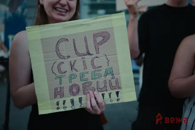 Smiling person holds a yellow cardboard sign with large handwritten letters; braces are visible, background blurred.