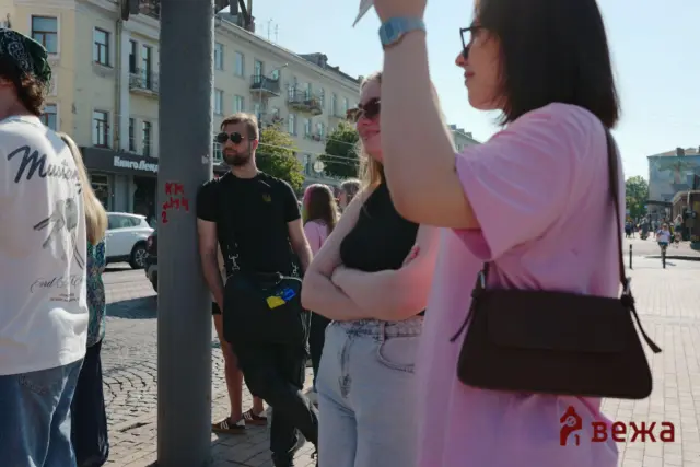 Group of people standing on a sunny city street, chatting near a lamppost with pastel buildings in the background