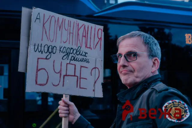A man wearing a dark jacket with patches holds a cardboard protest sign with handwritten Cyrillic text in red and black ink.