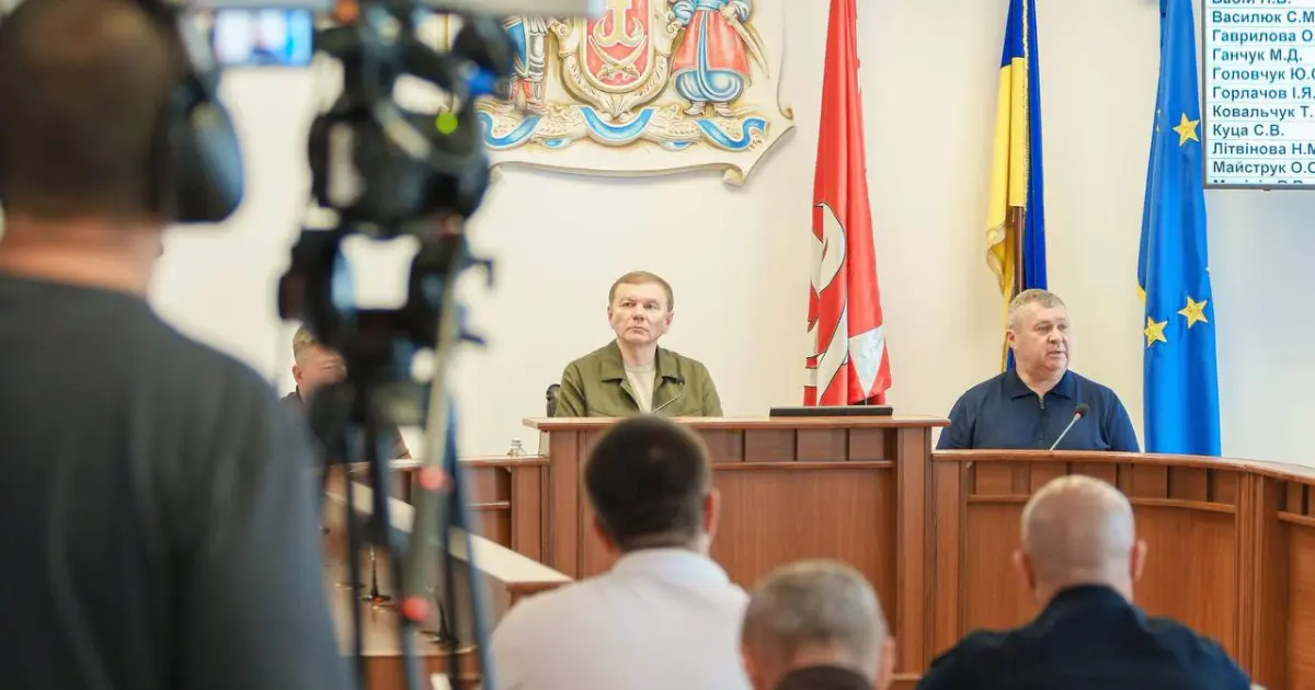 Press conference with two men at a wooden dais, Ukrainian and European Union flags behind, camera in foreground.