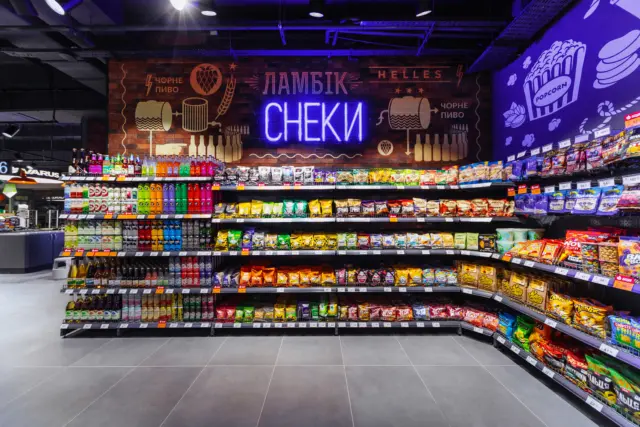 Supermarket aisle filled with colorful snack bags on shelves and a blue neon sign reading 'СНЕКИ' on a brick wall above them.