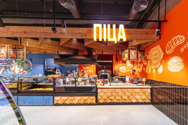 Front counter of a pizza shop with a glass display, staff in hairnets behind, and a warm orange wall with drawings.