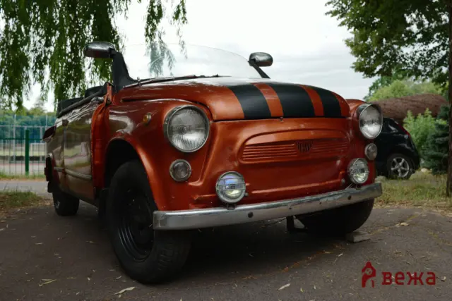 Front view of a red vintage convertible with black racing stripes parked under trees, showing round headlights and chrome bumper.