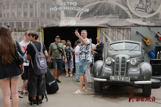 Group of people around a vintage car display; man takes a selfie with his phone in front of a gray classic vehicle outside a themed exhibit poster.