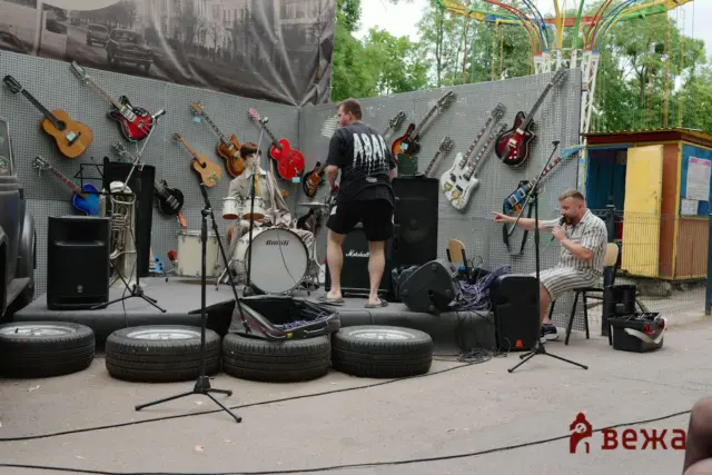 Outdoor stage setup with numerous guitars hanging on a perforated wall, a drum kit, and a man in a black shirt facing away toward a speaker system while another man sings into a microphone.