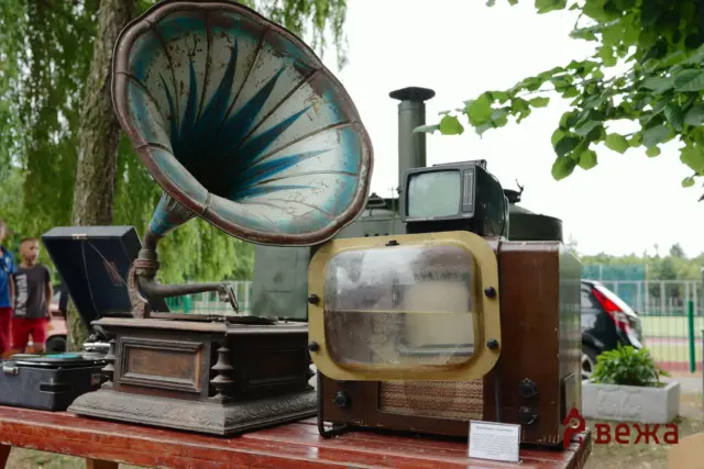 Old rusty gramophone with a large horn on a wooden table outdoors, with trees and people in the background.