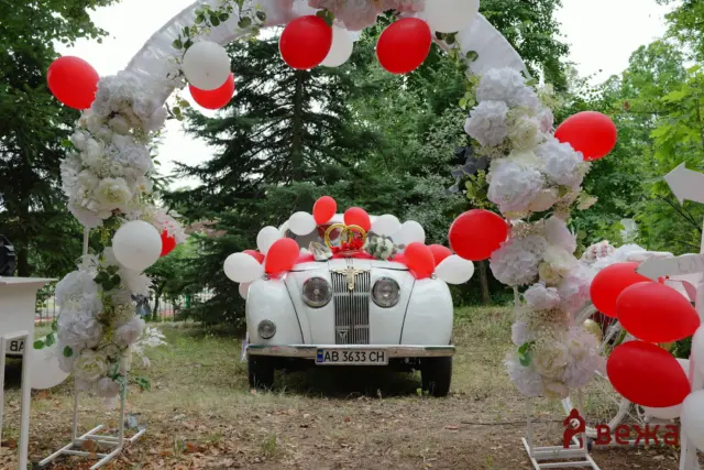 Vintage white car decorated with red and white balloons and floral arches for a wedding ceremony.