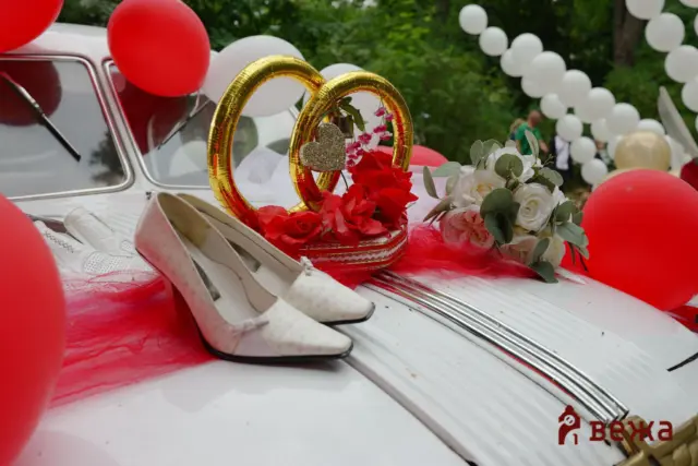 White vintage car trunk decorated for a wedding with red and white balloons, a gold heart wreath, red roses, and white shoes nearby outdoors.