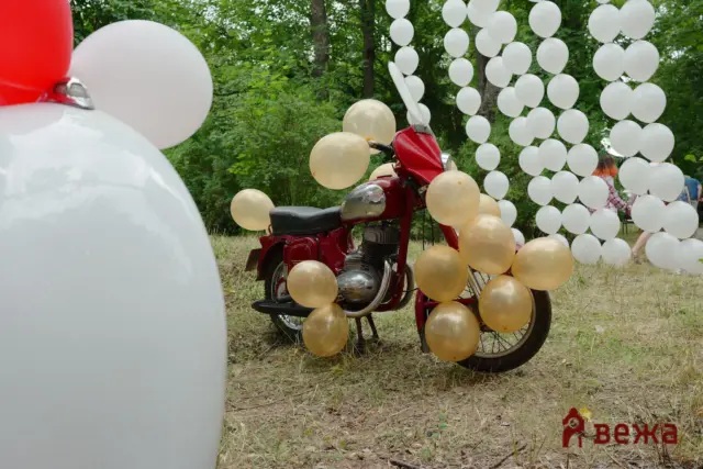 Red motorcycle decorated with gold and white balloons, outdoors among green trees with white balloon columns in the background.