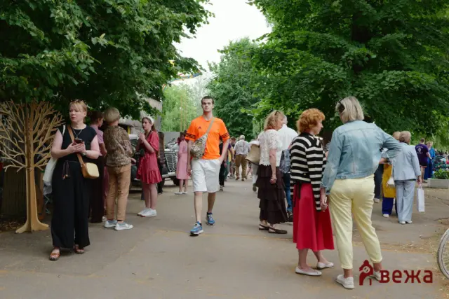 Crowd of people at an outdoor event on a tree-lined path; a man in a bright orange shirt stands center while others chat nearby.
