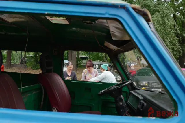 People stand outside a green vehicle, seen through an open window of a blue van, in a parking area with trees in the background.