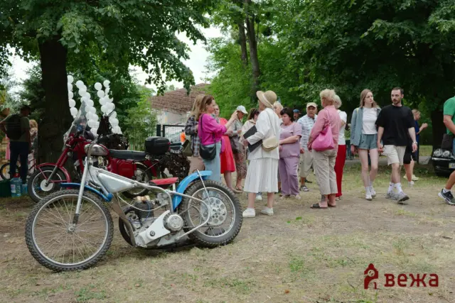 Group of people chatting at an outdoor event with vintage motorcycles parked in the foreground under trees.