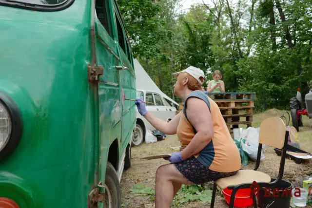 Person painting a green van outdoors with a brush, wearing an orange shirt, white cap, and purple gloves.