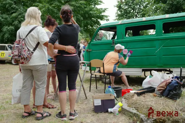 Group of people standing and talking near a green van in a park, with bags and supplies on the ground.