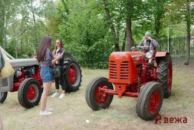 Two young women converse beside a red vintage tractor in a park-like yard with trees behind them.