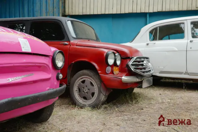 Three vintage cars parked bumper to bumper in an outdoor lot—pink, red, and white, with a blue wall in the background.