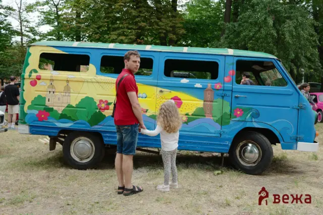 Man in a red shirt holds a child's hand beside a bright, mural-painted van in a park setting,