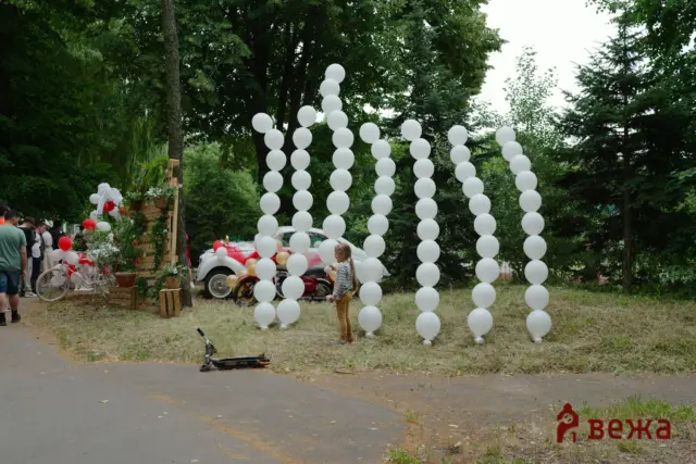 Outdoor party scene with tall white balloon columns arching over a path; people gather on the left near a decorated car.
