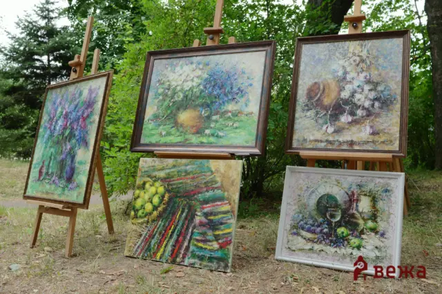 Outdoor art display with several framed paintings on wooden easels among green trees and grass, showcasing impressionist-style works.
