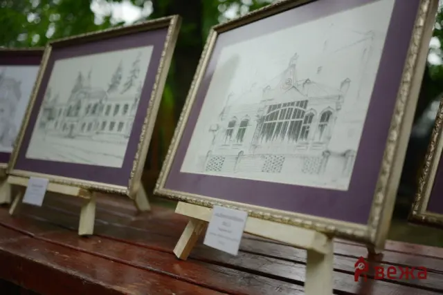 Row of framed architectural sketches on easels outdoors with purple mats and labels beneath, blurred trees in the background.