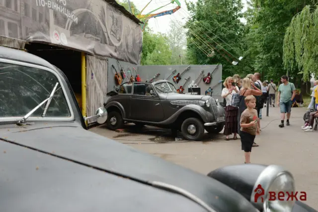 Vintage cars parked along a street with a wall displaying guitars; people walk and chat nearby on a sunny day.
