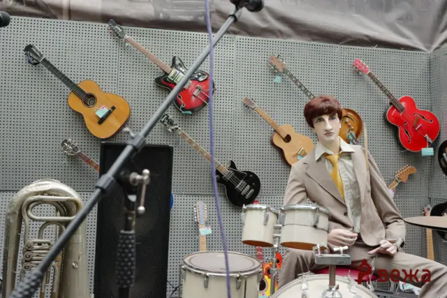 Mannequin in a beige suit and yellow tie seated at a drum kit in front of a wall of guitars in a music store display.