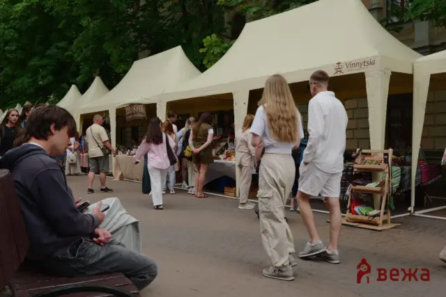 Outdoor market scene with white tents and shoppers browsing stalls; green trees in the background.