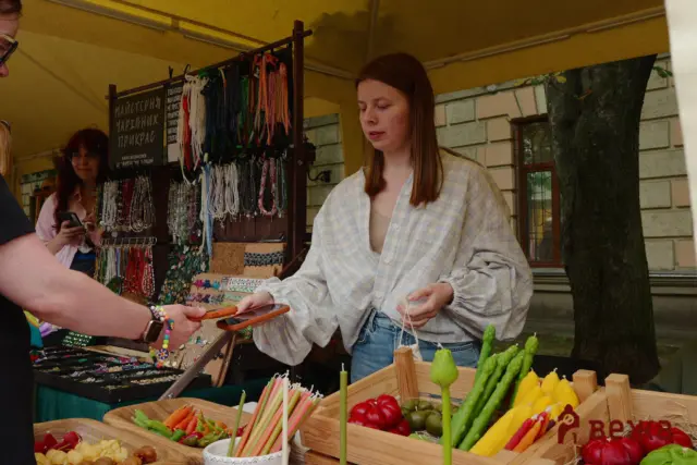 Market vendor hands a customer change or payment at an outdoor stall; colorful beaded jewelry and fresh produce (peppers, corn) on display.
