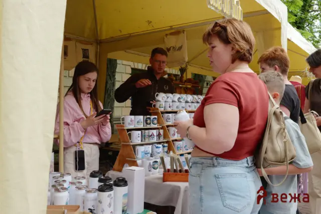 Group of shoppers at an outdoor market stall looking at ceramic mugs and tumblers in a display rack.