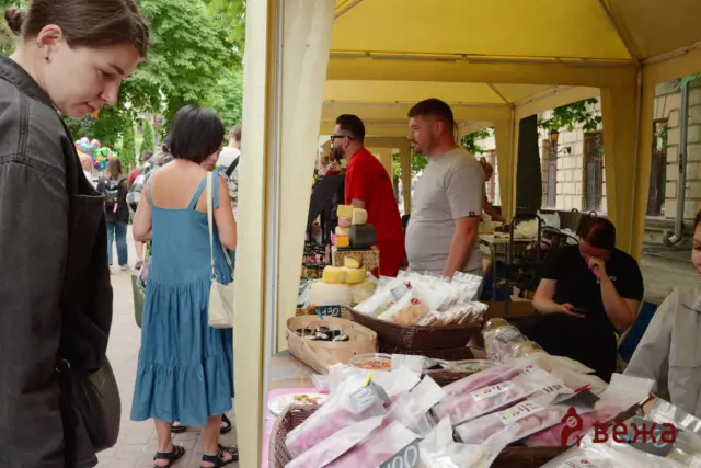 Outdoor market stall under a yellow canopy; a vendor in a gray shirt stands behind a table of cheeses and snacks while shoppers browse nearby, including a woman in a blue dress facing the display.