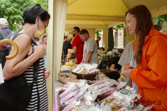 Two women at an outdoor market stall browsing packaged snacks and cheeses on a table under a cream canopy, with other shoppers in the background.