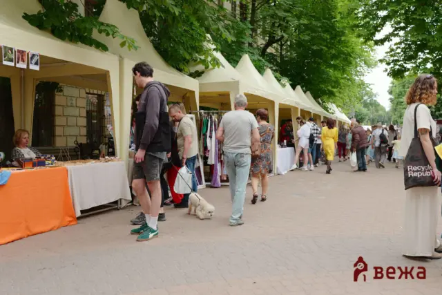 Outdoor market with beige canopy tents along a tree-lined street; shoppers browse clothing and crafts, a small white dog on a leash nearby.