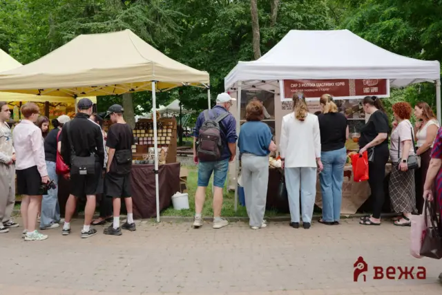 Group of people in a queue at an outdoor market with beige and white tents and vendor stalls behind them in a park setting.