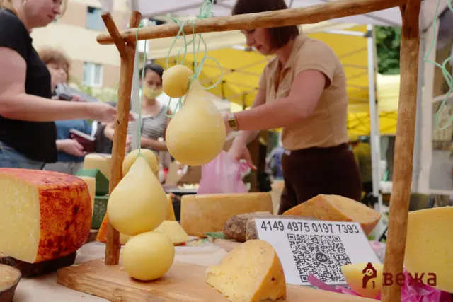 Close-up of yellow gourd-shaped cheeses hanging from a wooden frame at a market stall, with blurred shoppers in the background.