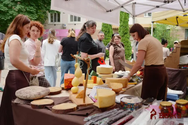 Outdoor market cheese stall with wheels and blocks of cheese on display; several people browse and chat nearby under a canopy.