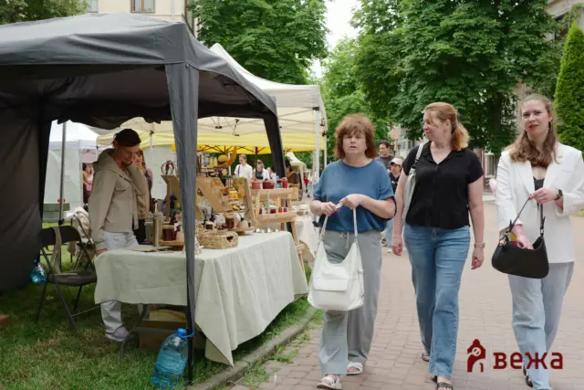 Outdoor street market with canopied stalls and wooden displays; four women walk past along a tree-lined sidewalk.