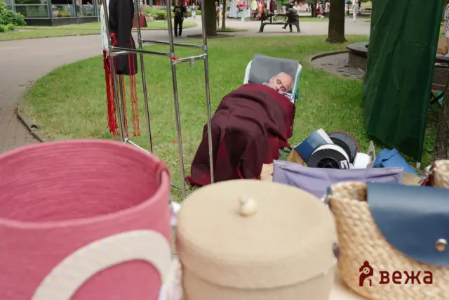 Older man sleeping in a reclining chair at an outdoor park event, covered with a burgundy blanket. Behind him, people walk near market stalls and trees.