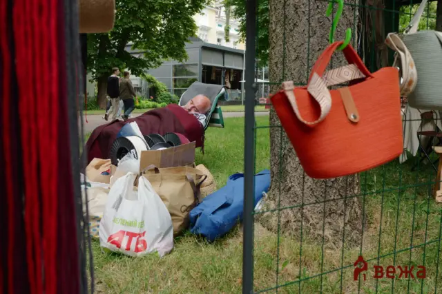 Older man sleeping on a folding lounge chair under a burgundy blanket, next to bags and fabric in an outdoor market setup.