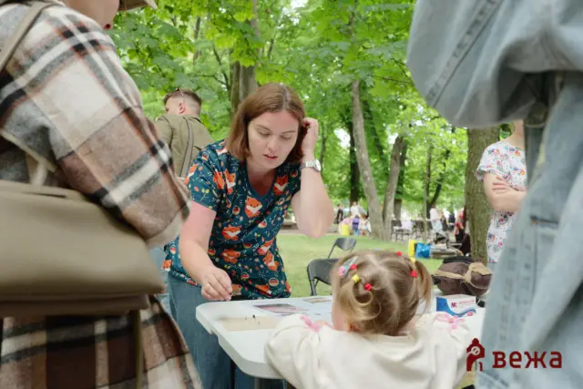 Woman in a blue fox-patterned blouse leans over a table at an outdoor park event, guiding children as they play or craft nearby.