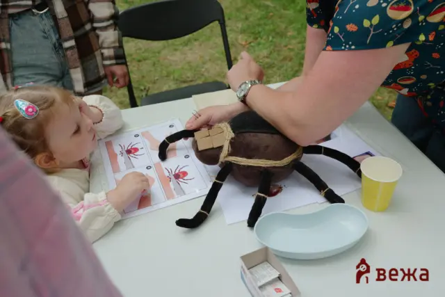 Child sits at an outdoor craft table, working on a spider-themed project with a black spider prop and worksheets nearby.
