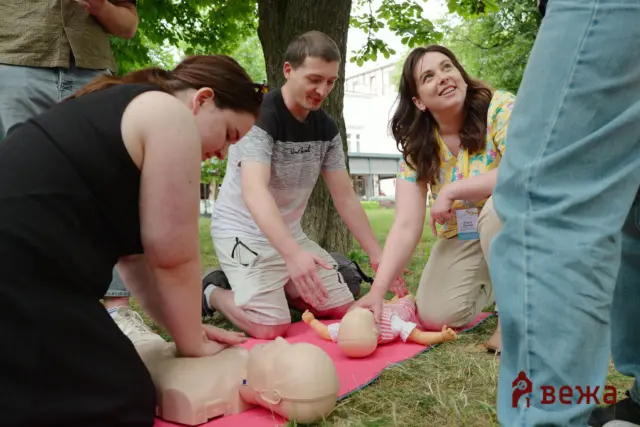Group of adults practicing CPR on an adult mannequin outdoors on a pink mat under a tree.