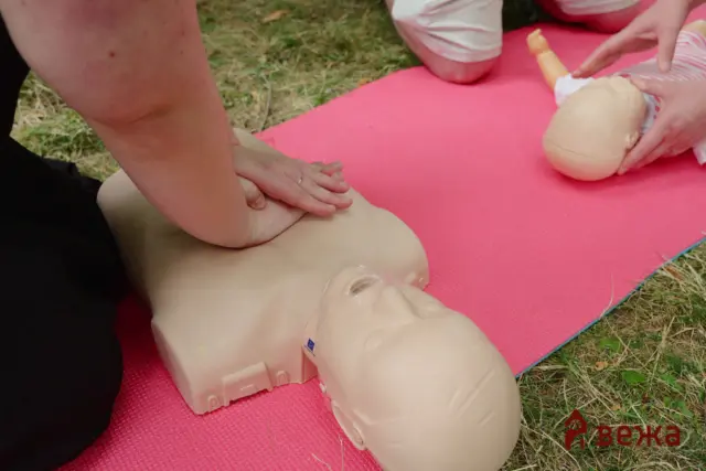 Close-up of hands performing CPR on an infant mannequin placed on a pink mat outdoors.