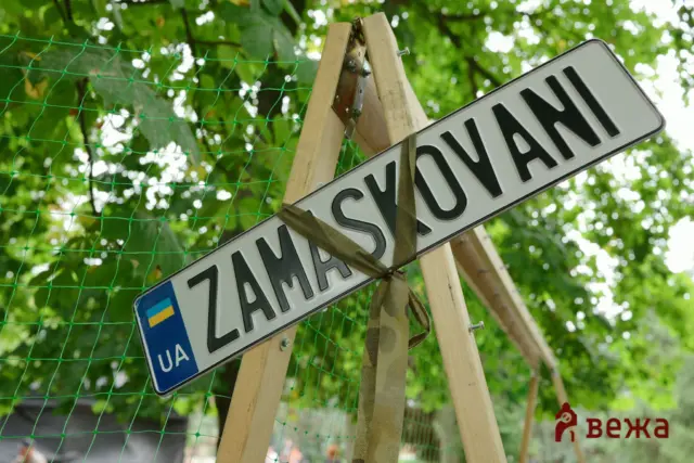 Diagonal street sign mounted on a wooden post, with green construction netting and lush trees in the background; small Ukrainian flag patch visible on the sign corner.